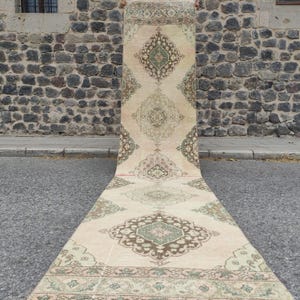 Puede incluir: Una alfombra de pasillo larga y estrecha con una base de color crema y patrones geométricos de medallones repetidos en tonos verde salvia, marrón y rosa suave. El diseño incluye motivos florales intrincados y un borde decorativo.