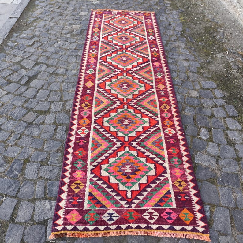 May include: A long, rectangular rug with a geometric pattern. The rug features a burgundy background with diamond and triangle shapes in shades of red, orange, green, and white. The rug is likely made of woven fabric.