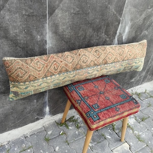 May include: A long, rectangular, patterned pillow in shades of brown, orange, and blue rests against a gray wall. Below it is a small, square stool with a red and blue geometric design and wooden legs. The items appear to be vintage or antique.