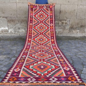 May include: A long, rectangular, hand-woven rug with a geometric pattern. The rug features a deep burgundy background with vibrant diamond and triangle shapes in orange, red, green, and white. The edges are finished with a fringe.
