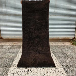 May include: A long, rectangular, dark brown rug with a shaggy texture. The rug is held up against a corrugated metal background and a tiled floor. The rug appears to be made of a soft, plush material.