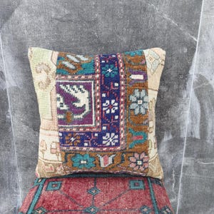Puede incluir: Cojín decorativo cuadrado con diseño de patchwork. El cojín presenta una mezcla de colores, incluyendo beige, turquesa, morado y azul marino. El cojín está colocado sobre una alfombra roja estampada.