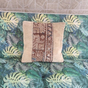 Op de afbeelding: Een beige kussen met een bruine en blauwe gepatroneerde stoffen inzet. Het kussen ligt op een bank met een groen en wit tropisch bladpatroon.