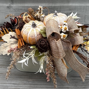 May include: A decorative centrepiece featuring two small pumpkins, one orange and one white, nestled in a metal container. The arrangement includes hessian ribbon, cinnamon sticks, dried leaves, twigs, and small white flowers with yellow centres.