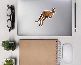 May include: A brown kangaroo sticker on a silver laptop computer. The laptop is on a white desk with a notebook, a pencil, and a mouse.