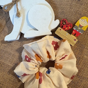 Puede incluir: Una scrunchie crema con un estampado floral en naranja, rosa y azul. Un conejito de madera blanco con un lazo de cuerda está al lado de la scrunchie. Corazones de madera decorativos y una figura de niña también están en la imagen.