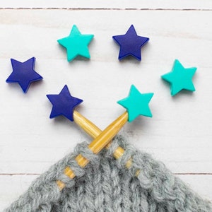 May include: Two wooden knitting needles with blue and turquoise star-shaped stitch markers. The needles are crossed and resting on a grey knitted fabric.