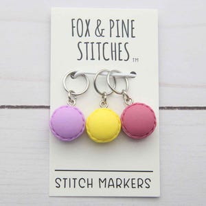 Könnte beinhalten: Drei Maschenmarkierer in Form von Makronen in Lila, Gelb und Rosa, die an einer weißen Karte hängen. Auf der Karte steht "FOX & PINE STITCHES" und "STITCH MARKERS". Die Marker sind an silberfarbenen Ringen befestigt.