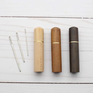 May include: Three cylindrical wooden needle cases in light, medium, and dark brown tones, each featuring a thin gold-colored metal band around the center. Three silver-colored metal sewing needles of varying lengths are positioned to the left.