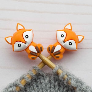Könnte beinhalten: Zwei orangefarbene Stricknadelspitzen in Form von Füchsen auf einem Paar Holzstricknadeln mit grauem Garn.