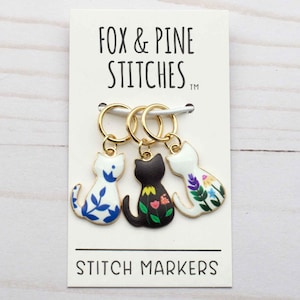 May include: Three gold stitch markers with cat charms. The cats are white with blue floral designs, black with red and green flowers, and white with purple and yellow flowers. The card reads "Fox & Pine Stitches" and "Stitch Markers".