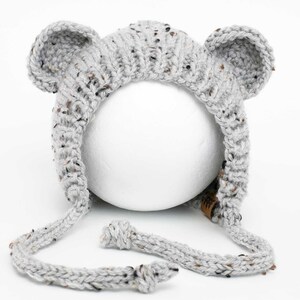 Peut inclure: Un bonnet bébé en tricot gris avec des oreilles d'ours et une longue cravate. Le bonnet est fait d'un fil doux et texturé.