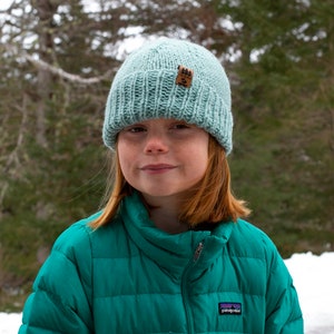 Op de afbeelding: Een jong persoon draagt een tealblauwe pufferjas met een lichtblauwe gebreide beanie met een bruine patch. De beanie heeft een geribbelde textuur. De persoon staat voor een besneeuwde achtergrond.