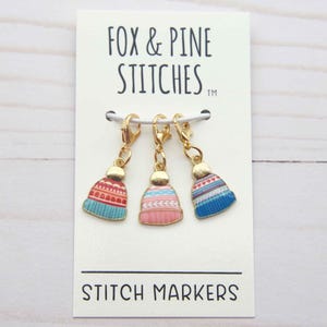 Könnte beinhalten: Drei goldfarbene Maschenmarkierer in Form von Miniatur-Strickmützen, jeweils mit unterschiedlichen Farbschemata: Rot und Blau, Rosa und Blau. Die Marker hängen an einer weißen Karte mit dem Text "FOX & PINE STITCHES" und "STITCH MARKERS".