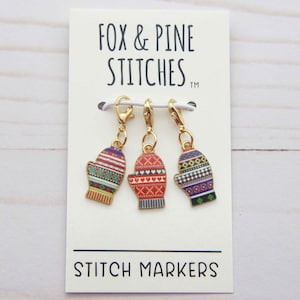 Op de afbeelding: Drie stekenmarkeerders in de vorm van wanten met kleurrijke patronen, gepresenteerd op een witte kaart. De wanten hebben patronen in rood, blauw en groen. De kaart leest "FOX & PINE STITCHES" en "STITCH MARKERS".