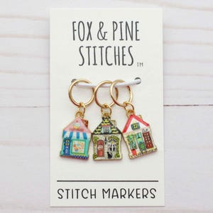 Puede incluir: Tres marcadores de punto con forma de casa con anillos dorados se muestran en una tarjeta blanca. Los marcadores están pintados en varios colores, incluyendo rosa, verde y rojo, con detalles intrincados. La tarjeta dice "FOX & PINE STITCHES" y "STITCH MARKERS."