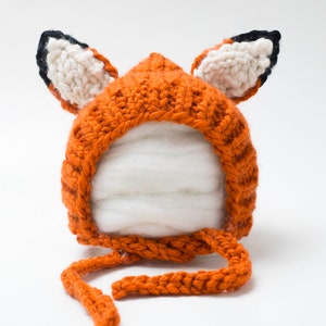 May include: A knitted fox hat for a baby, with orange and white fur and black ears. The hat has a long tie that can be tied under the chin.