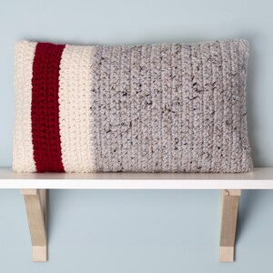 May include: A grey, white and red striped crocheted cushion on a white shelf with wooden brackets.