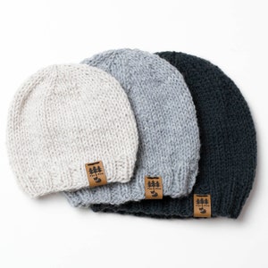 May include: Three knitted beanies in different colors: white, gray, and black. Each beanie has a brown leather tag with the text "Fox & Pine" and a small tree graphic.
