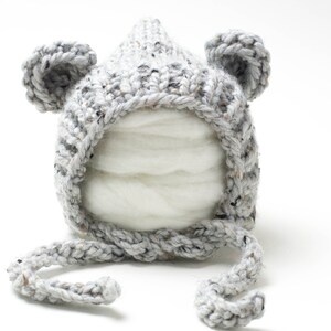 Peut inclure: Bonnet bébé en tricot gris avec des oreilles d'ours et une longue cravate. Le bonnet est fait d'un fil doux et texturé.