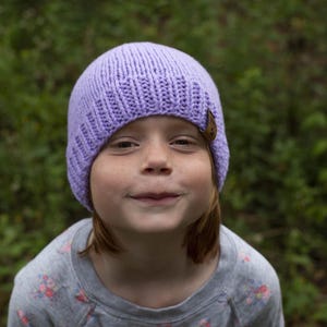 Op de afbeelding: Een jong persoon draagt ​​een paarse gebreide beanie met een bruine knoop aan de zijkant. De beanie is over het voorhoofd getrokken en de persoon kijkt naar de camera.