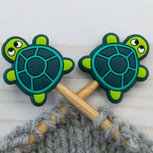 May include: Two wooden knitting needles with green and blue turtle-shaped stitch markers. The needles are resting on a gray knitted fabric.