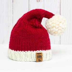 Puede incluir: Sombrero de Papá Noel de punto rojo y blanco con un pompón blanco en la parte superior. El sombrero tiene una etiqueta de cuero marrón con el texto "Tree & Vine" en ella.