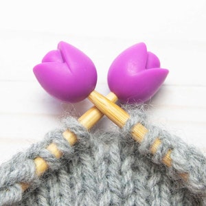 May include: Two wooden knitting needles with purple tulip-shaped ends, crossed over each other, with grey yarn on the needles.
