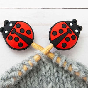 May include: Two wooden knitting needles with ladybug-shaped toppers. Each topper is a round, black and red ladybug with white eyes and black spots. The needles are crossed and inserted into a piece of gray, chunky knit fabric.