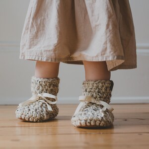 May include: Close-up of a child's feet wearing handmade crochet booties. The booties are a neutral color with a textured design and cream-colored ribbon ties. The child is wearing a beige dress. The image is taken indoors on a wooden floor.