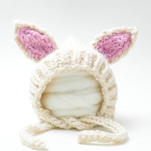 Puede incluir: Un gorro de bebé de punto blanco con orejas de conejo rosas. El gorro tiene una corbata que se puede atar debajo de la barbilla.