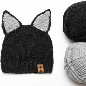 May include: A black knitted hat with gray cat ears and a brown leather tag that says "Fox & Pine". The hat is next to two balls of yarn, one black and one gray.
