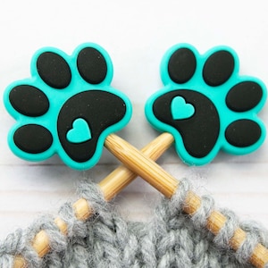 Op de afbeelding: Twee turquoise blauwe steekmarkeerders in de vorm van een poot met zwarte voetkussens en een klein turquoise hartje op elke markeerder. De markeerders zitten op houten breinaalden.