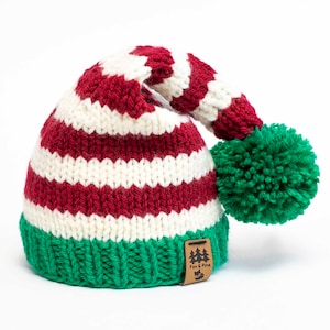Puede incluir: Gorro de elfo de punto a rayas rojas y blancas con un pompón verde y una etiqueta de cuero marrón que dice "Fox & Pine".