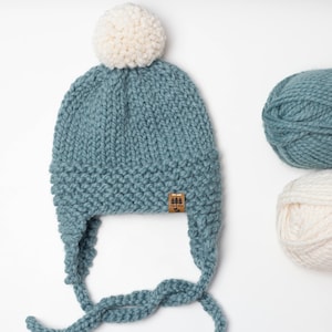 May include: A blue knitted hat with a white pom pom and ear flaps. The hat is tied with a long blue string. Two balls of yarn are shown in the background, one blue and one white.