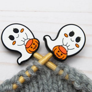 May include: Two wooden knitting needles with ghost-shaped stitch markers holding small orange pumpkins. The needles are crossed and resting on a grey knitted fabric.