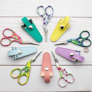 May include: Eight pairs of small, colourful scissors with floral patterns, each with a matching leather case. The scissors are arranged in a circle on a white wooden surface.