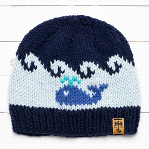 May include: A blue knitted hat with a white whale and waves design. The hat has a navy blue brim and a brown leather tag with the text "Fox & Pine" on it.