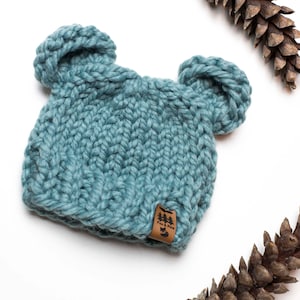 May include: A light blue knitted hat with two ear flaps, resembling bear ears. The hat has a brown leather tag with the text "Fox & Fern" on it.