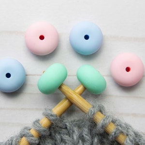 May include: Four colourful round wooden beads with holes in the centre, two light blue, one pink, and one green, are strung on two wooden knitting needles with grey yarn.