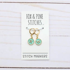 May include: Two gold stitch markers with turquoise round charms featuring the letters "R" and "W". The markers are on a white card with the text "Fox & Pine Stitches" and "Stitch Markers".