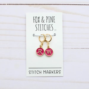 Puede incluir: Dos marcadores de punto de color rosa con aros dorados y las letras "R" y "W" en ellos. Los marcadores están en una tarjeta blanca con el texto "Fox & Pine Stitches" y "Stitch Markers".