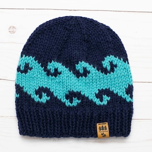 May include: A navy blue knitted beanie with a teal wave pattern. The beanie has a small brown label with the text "Fox & Pine" on it.