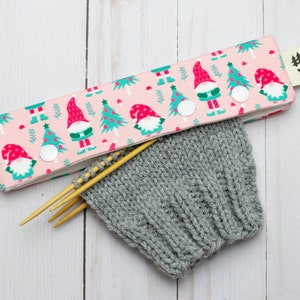 May include: A pink knitting needle case with a pattern of gnomes and Christmas trees, secured with white snaps. A gray knitted hat and wooden knitting needles are visible.