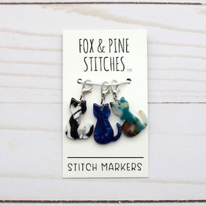 Op de afbeelding: Drie kattenvormige steekmarkeerders op een witte kaart met de tekst "Fox & Pine Stitches TM" en "Stitch Markers". De markeerders zijn gemaakt van doorschijnend plastic en hebben een zwart-witte kat, een blauwe kat en een groene en bruine kat.
