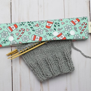 Puede incluir: Un protector de agujas de tela verde y blanco con un estampado navideño de tazas de café, bastones de caramelo y copos de nieve. El protector de agujas está unido a un par de agujas de tejer de madera y a un proyecto de tejido de color gris.