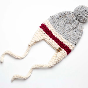 May include: A gray and white knit hat with a burgundy stripe and a fluffy pom pom on top. The hat has ear flaps that tie under the chin.