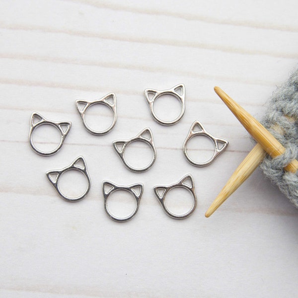 Op de afbeelding: Zilverkleurige steekmarkeerders in de vorm van een kat op een wit houten oppervlak. Breinaalden zijn in een grijze gebreide stof gestoken. De markeerders zijn klein, met een cirkelvormig lichaam en kattenoordetails.