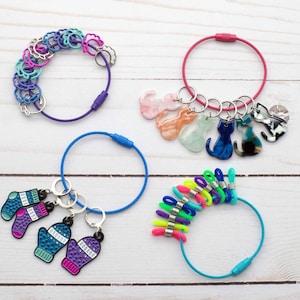 Pode incluir: Quatro conjuntos de marcadores de ponto coloridos para tricô ou crochê. O primeiro conjunto é um anel de marcadores roxo, rosa e azul. O segundo conjunto é um anel rosa com cinco marcadores em forma de gato. O terceiro conjunto é um anel azul com três marcadores em forma de luva. O quarto conjunto é um anel turquesa com oito marcadores coloridos.
