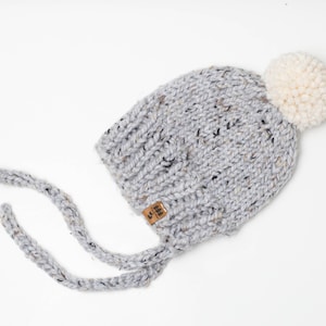 Könnte beinhalten: Eine graue gestrickte Baby-Mütze mit einem weißen Bommel oben. Die Mütze hat zwei lange Bänder, die unter dem Kinn gebunden werden können. Die Mütze hat ein kleines braunes Etikett mit dem Text "Little & Brave" darauf.